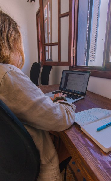 Woman focused on writing and working on a laptop at a wooden desk near an open window in a quiet workspace.