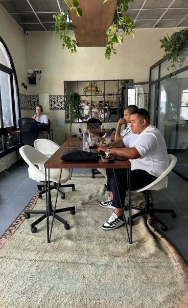 Two men seated on office chairs working on laptops at a long wooden desk over a rug in Noqta Space coworking interior.