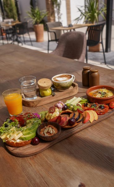 Gourmet breakfast platter with fresh fruit, avocado bagel toasts, cappuccino, orange juice, and a warm side dish on a wooden outdoor table.