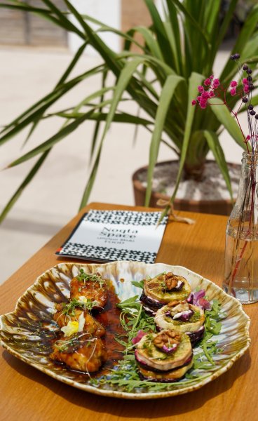 Plated dish featuring glazed chicken bites and goat cheese stack appetizers on a ceramic platter at a table with a Noqta Space menu and tropical plant background.