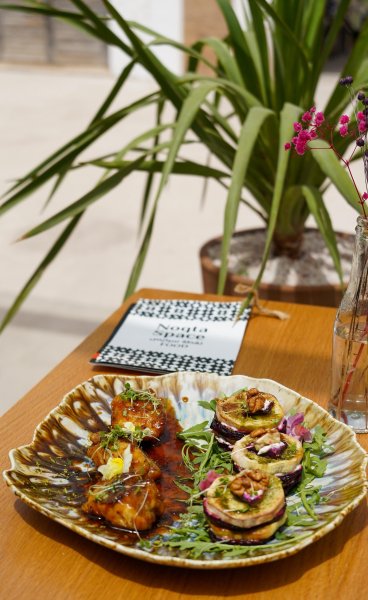 Plated dish featuring glazed chicken bites and goat cheese stack appetizers on a ceramic platter at a table with a Noqta Space menu and tropical plant background.